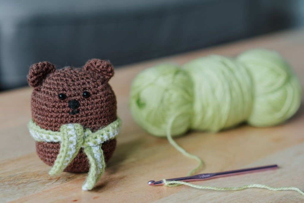 pexels photo 4792086 Closeup of small dark brown crochet toy bear and crochet next to light green threads on wooden table in bright room on blurred background